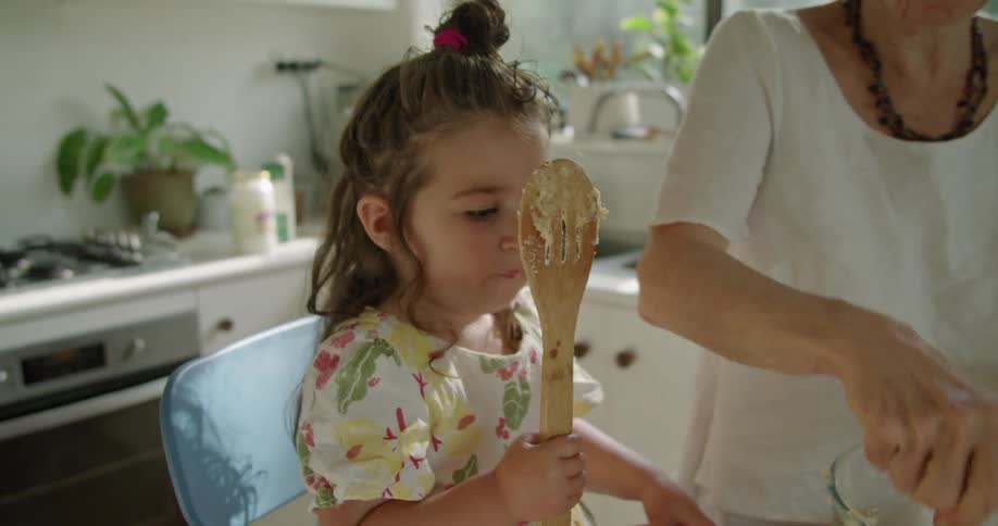 A little girl eats dough off of a wooden spool as she sits next to her grandmother who is baking in the kitchen