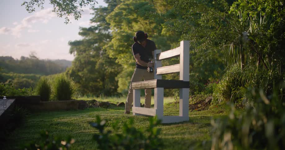 A man stands in a grassy field and puts on headphones and then starts to drill on a wooden bench