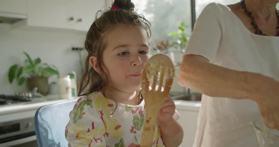 A young girl is in the kitchen with her grandmother licking a big wooden spoon