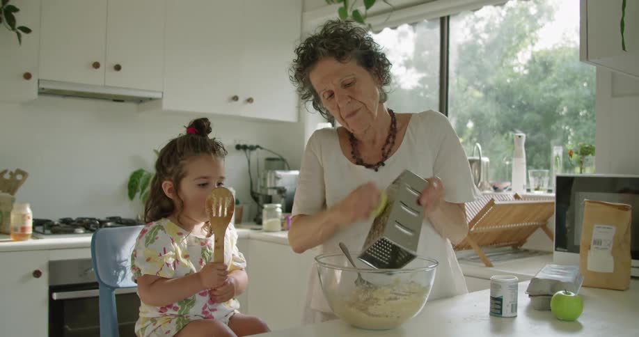 A young girl licks a mixing spoon while her grandmother attempts to show her how to grate a lemon in home kitchen during the day