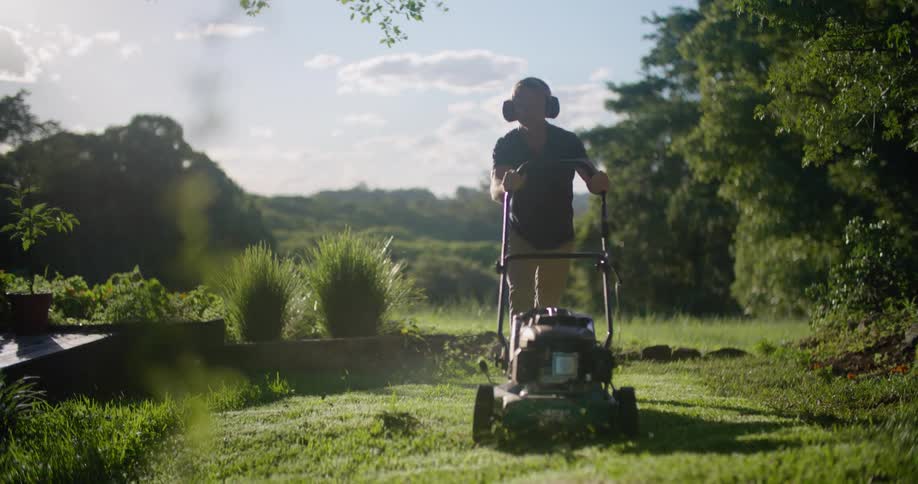 Man with headphones black shirt and brown pants mows the grass with lawn mower next to country house in the countryside