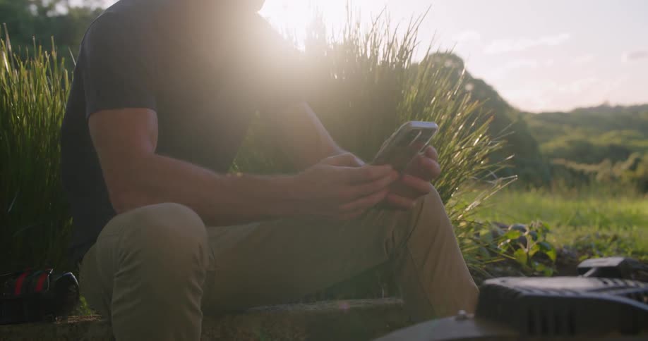 A man sits outside in a field and types on his cell phone
