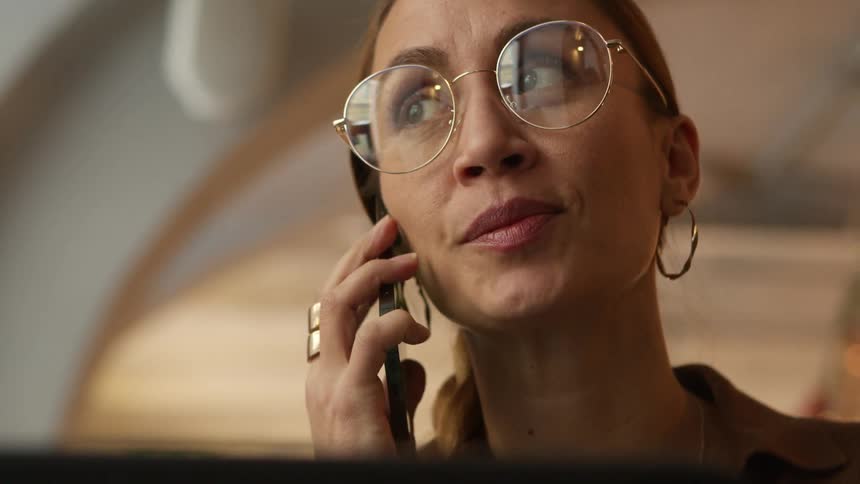 Woman with brown hair, glasses and hoop earrings makes phone call on smartphone in cafe