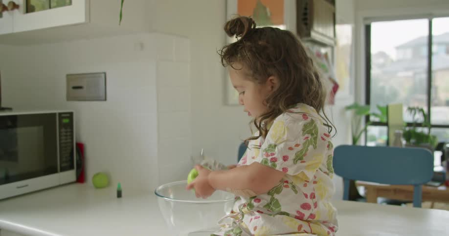 A little girl sits on a kitchen counter and holds an apple over a bowl as she tries to peel it