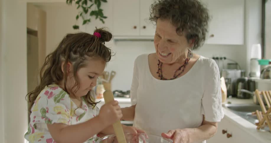 A little girl sits on a kitchen counter stirring a mixture in the bowl as her grandmother holds the bowl and leans down and talks