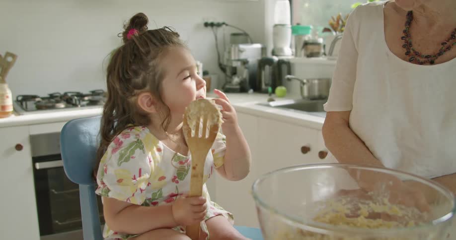 A little girl sits in the kitchen with her grandma and licks a spoon with batter on it and her grandma talks with her and claps