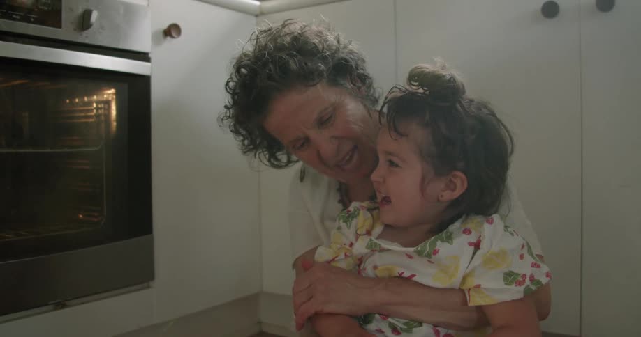 An elderly woman sits on the floor in the kitchen with her grand daughter in her lap and they clap hands together and laugh