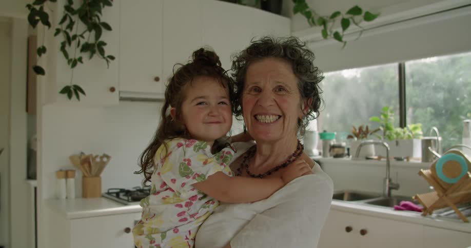 Grandmother hugs granddaughter in kitchen both smile until the girl sticks out her tongue