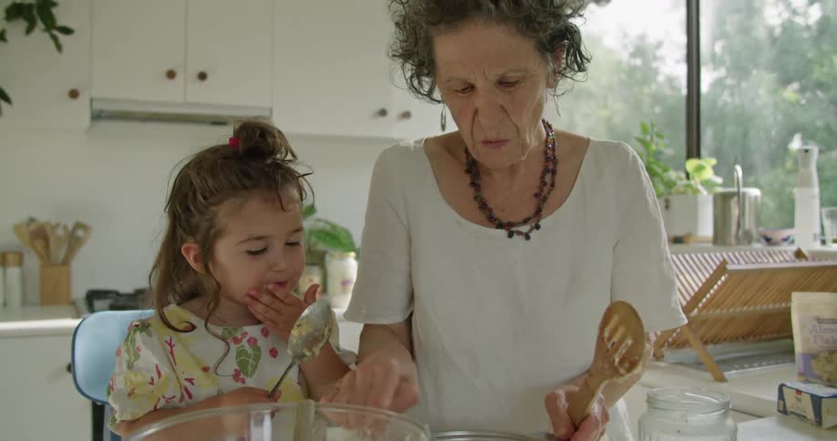 Grandmother with earrings necklace white t shirt makes food next to granddaughter tasting the food with spoon in the kitchen
