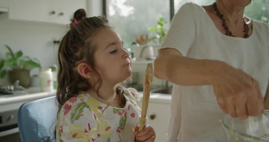 A grandmother cooks while her granddaughter licks the spoon