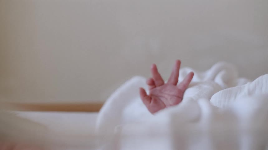 An infant in a cradle with white blankets holds his hand up