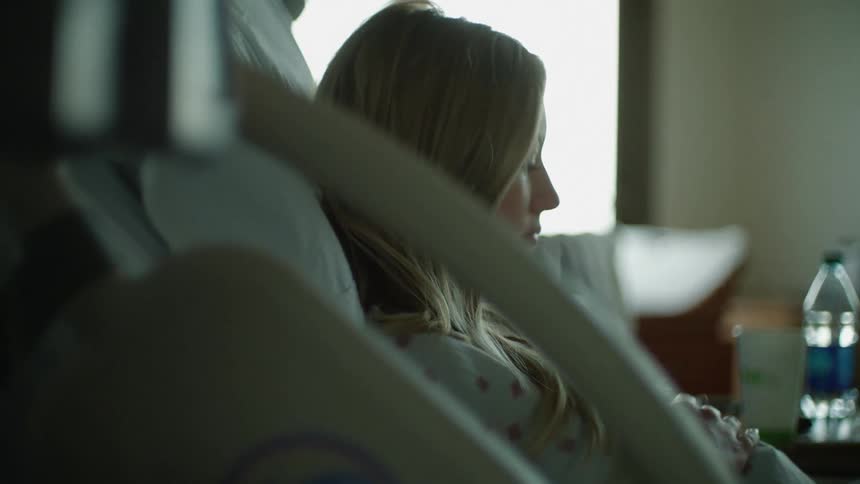 A pregnant woman sitting up in a hospital bed looks down as she rubs her belly