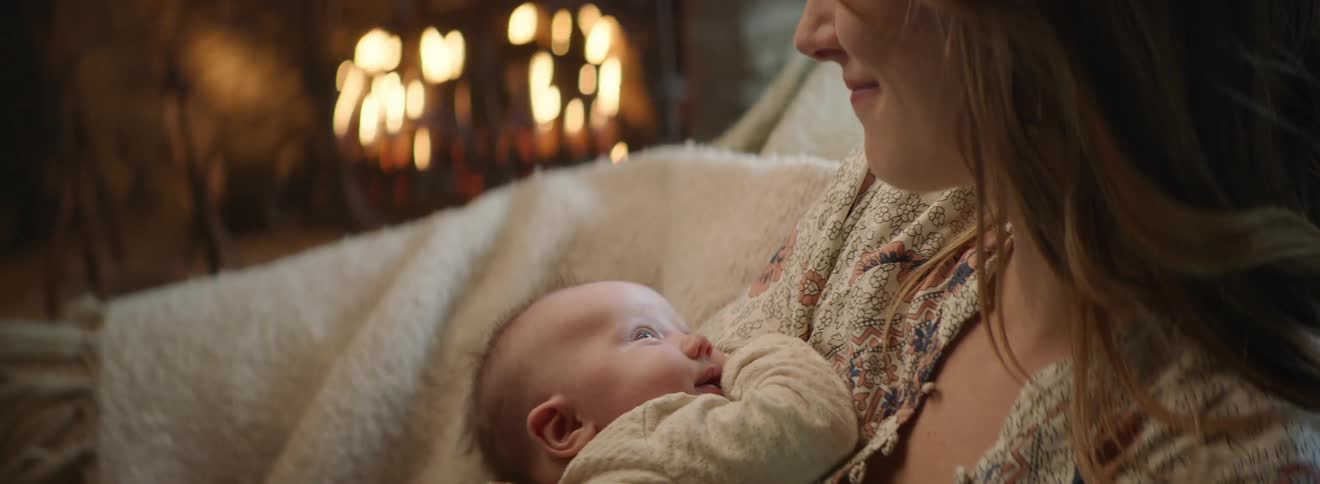 A mom looks down and her baby looks up at her as she snuggles and cradles him by the fire