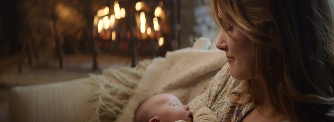 A mother smiles while holding her sleeping baby as they sit near a roaring fire