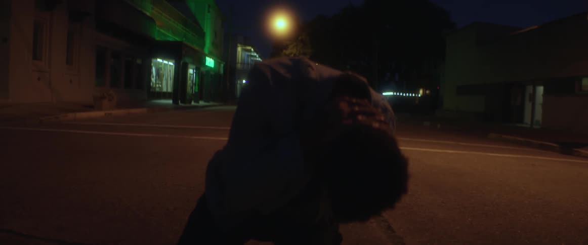 A man sits in a street next to dark buildings and puts his head in his hands and looks down at the ground and then up to the sky