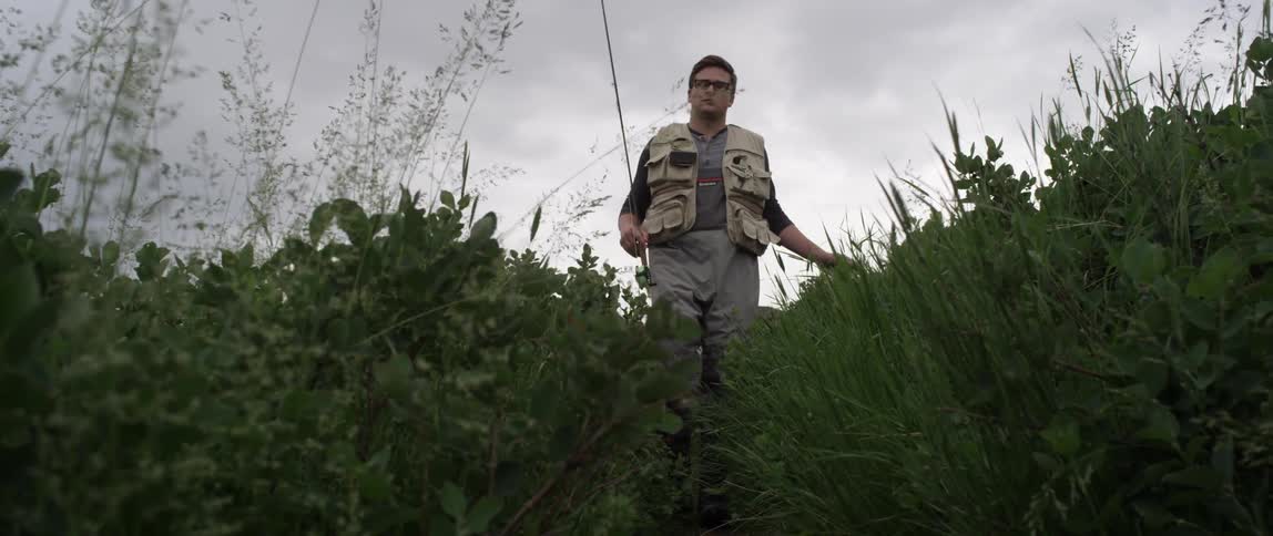 A man carrying a fishing pole walks through tall grass