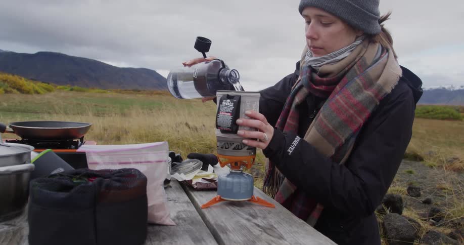 A woman pours water into a pitcher on a torch near a campsite