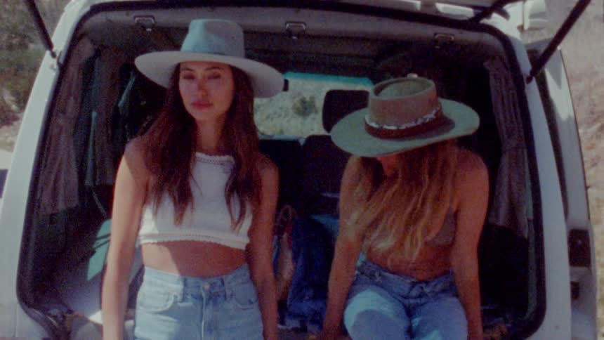 Two teenage girls wearing hats are sitting on the tailgate of a van