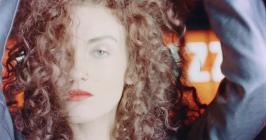 Woman with makeup black leather jacket holds her curly hair and dances in front of billboard