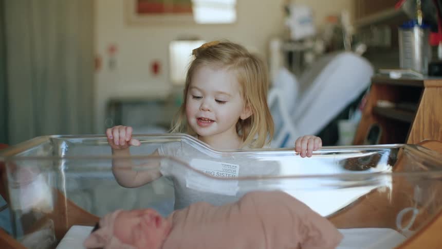 Big sister looking at a newborn baby resting on a transparent crib inside a nursery