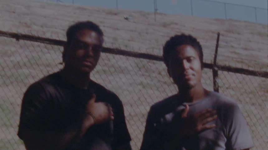 Two men are standing outside by a fence and holding their right hands against their chest