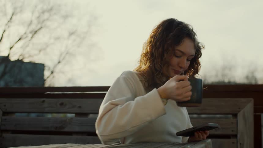 Woman with curly hair and white sweater drinks coffee from mug and uses smartphone outdoors sitting at table