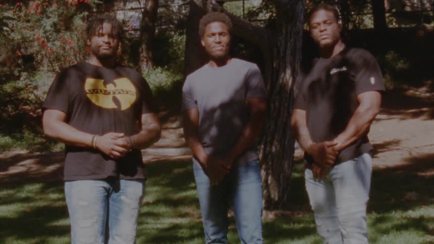 Men stand outdoors near tree posing with their hands clasped in front