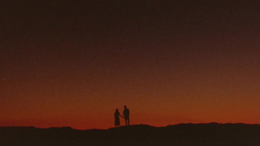 Man and woman holding hands are silhouetted against the red sky at dusk