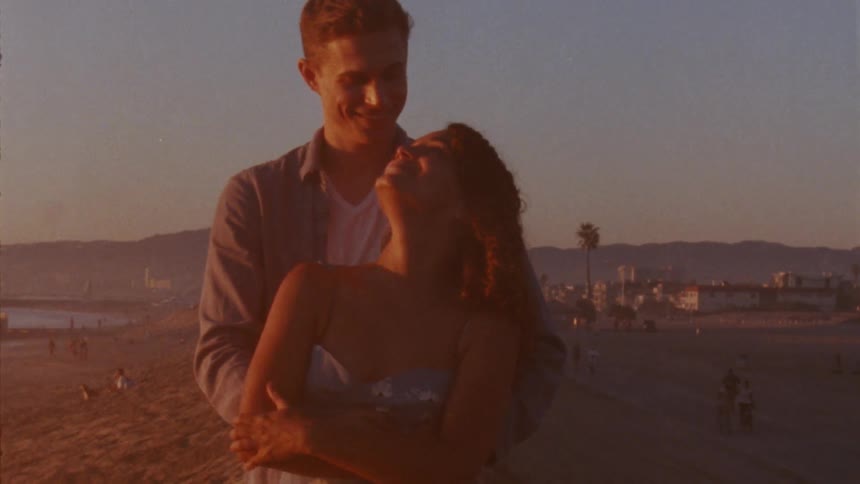 A woman smiles into the camera while standing on the beach until a man hugs her from behind