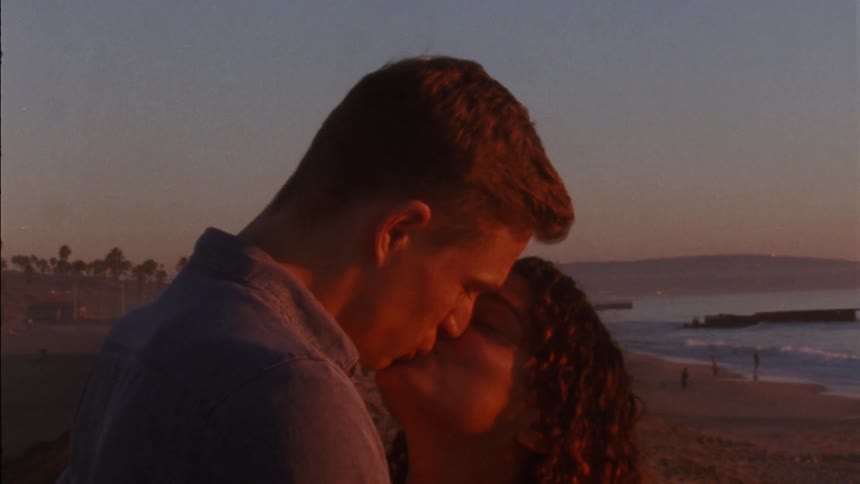 Man with blue shirt and woman with curly hair make out on the coast at sunset
