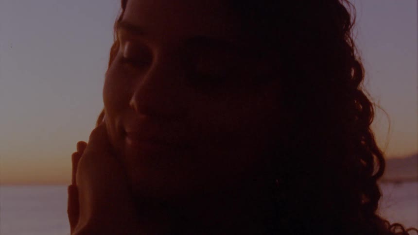 Woman back lit against red tinted horizon and water smiles as someone caresses her cheek