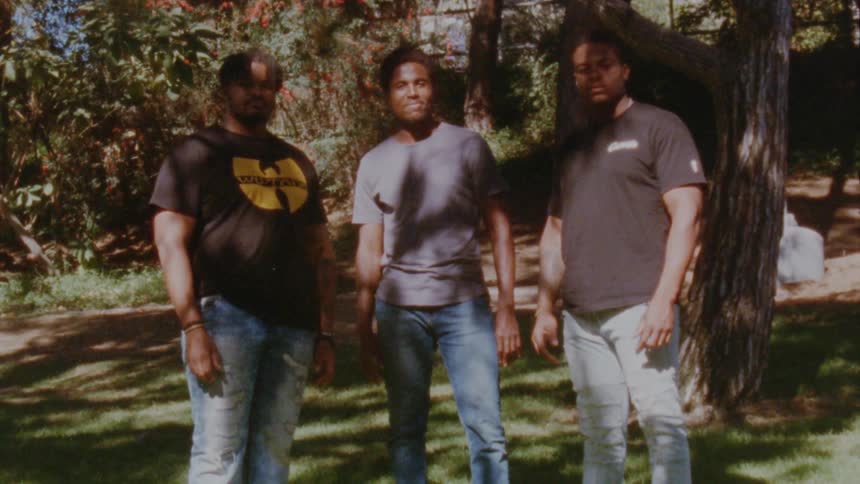 Three black guys are standing under a tree looking serious