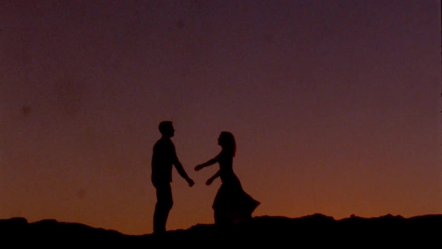The silhouette of a woman approaching and kissing a man outdoors at sunset
