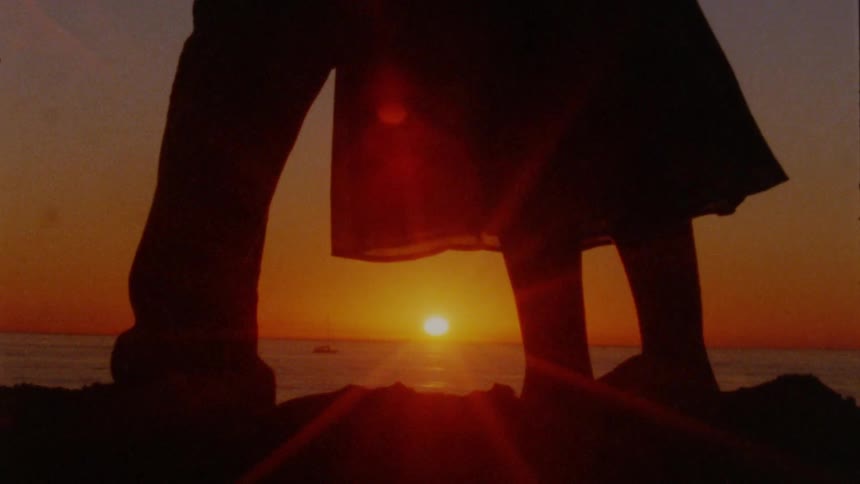 Close up of the legs of a man and woman standing closely together on the shore as the sun sets