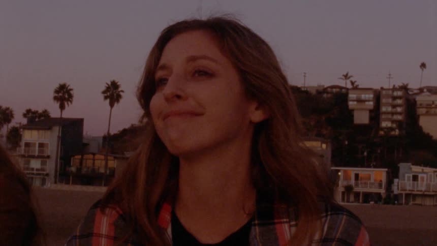 Woman with brown wavy hair and plaid shirt looks at her friend and smiles on the beach at sunset