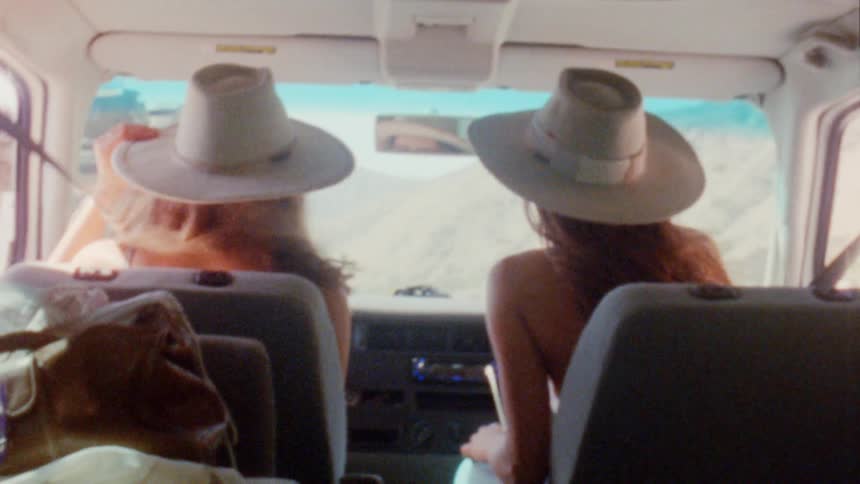 Two women with with brown wavy hair hats and sunglasses travel by van