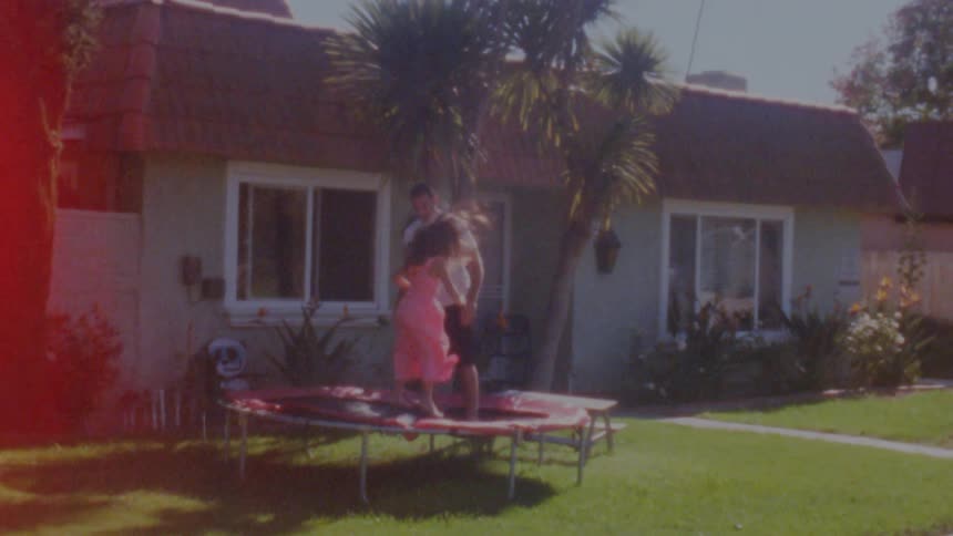 Father with beard long hair white t shirt and black shorts jumps on trampoline with daughter wearing pink dress in the garden