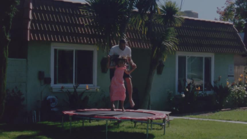 A man holds his daughters hands as they jump on a trampoline together outside a house