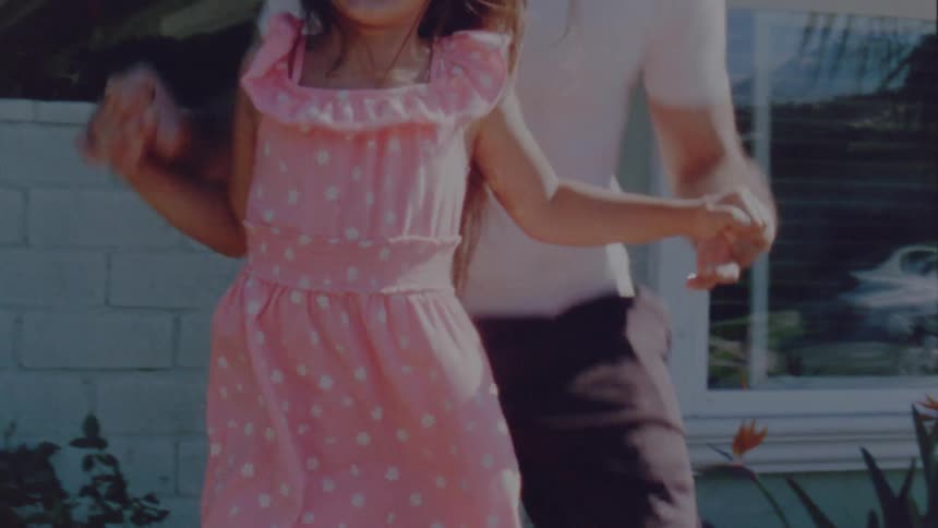 Father with beard long hair white t shirt and black shorts jumps on trampoline with daughter wearing pink dress in the garden