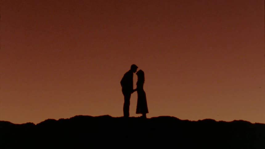 Man and woman holding hands and making out are silhouetted against the red sky