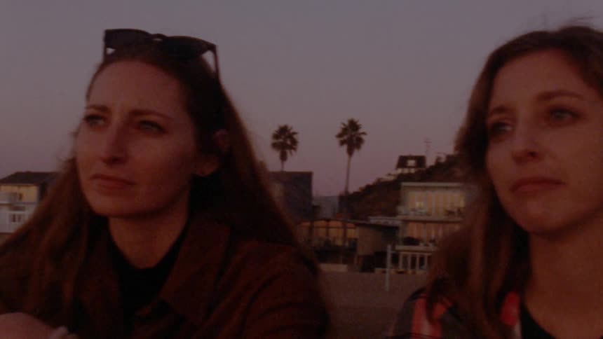 Two woman are sitting and smiling on a beach near an ocean front town at dusk