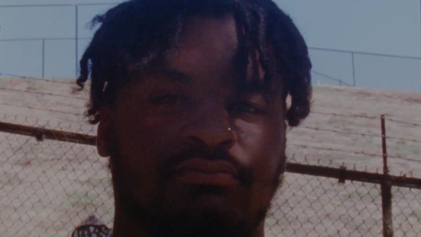Man with braids and beard stares at camera in front of wire netting