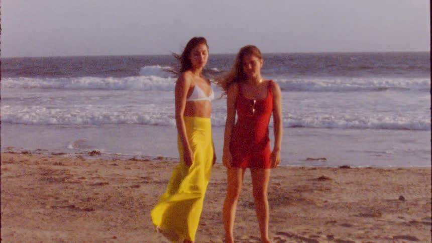 Woman with yellow skirt white bra stands next to woman with brown wavy hair and red dress on beach next to ocean