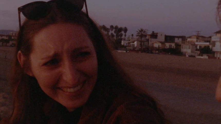 Two women sit on beach and laugh together as a jogger passes in background
