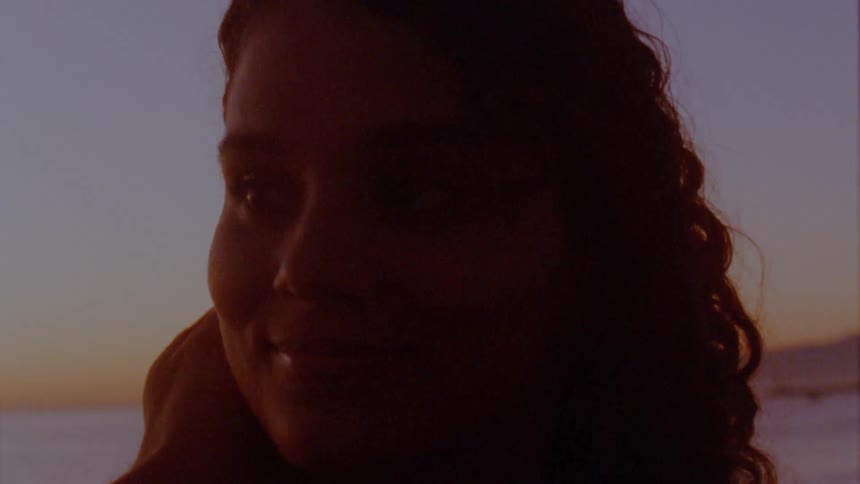 A woman smiles as a hand caresses her cheek on a beach at dusk