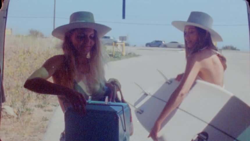 Two women grab a cooler and a surfboard out of the back of their van