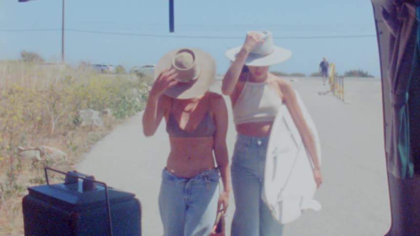 Vintage footage of two women one carrying a surfboard walking towards a vehicle and both of them taking off their hats and putting them in the vehicle