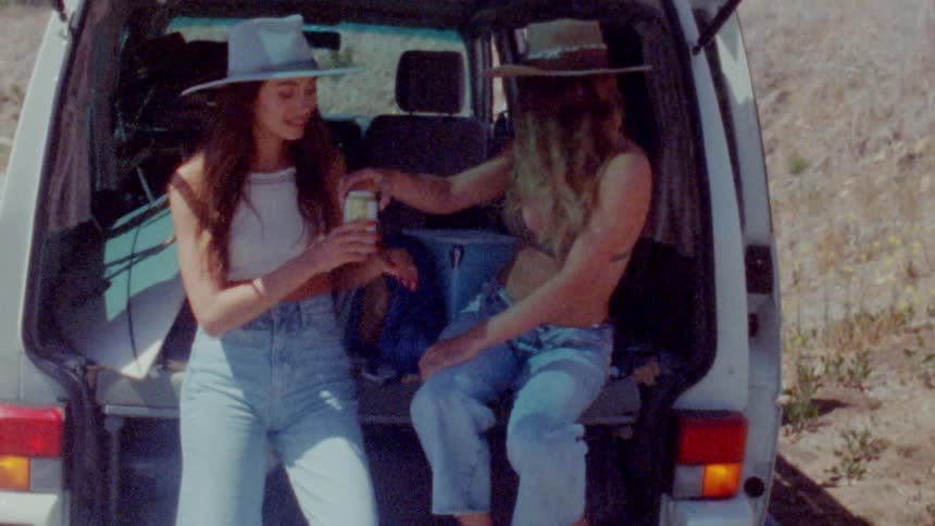 Two women are sitting on a parked car and drinking a beverage