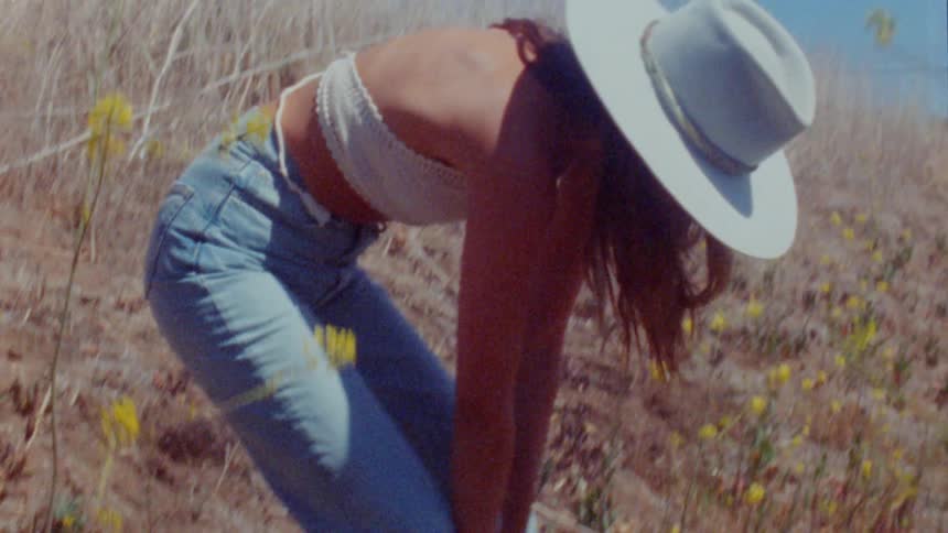 A woman stands up from crouching on the side of a hill filled with weeds and yellow flowers
