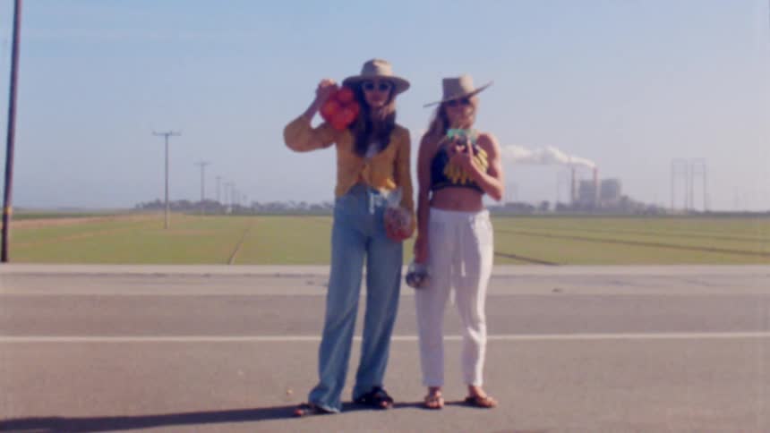 Two women stand holding produce as an orange rolls across the ground and car passes along road behind them